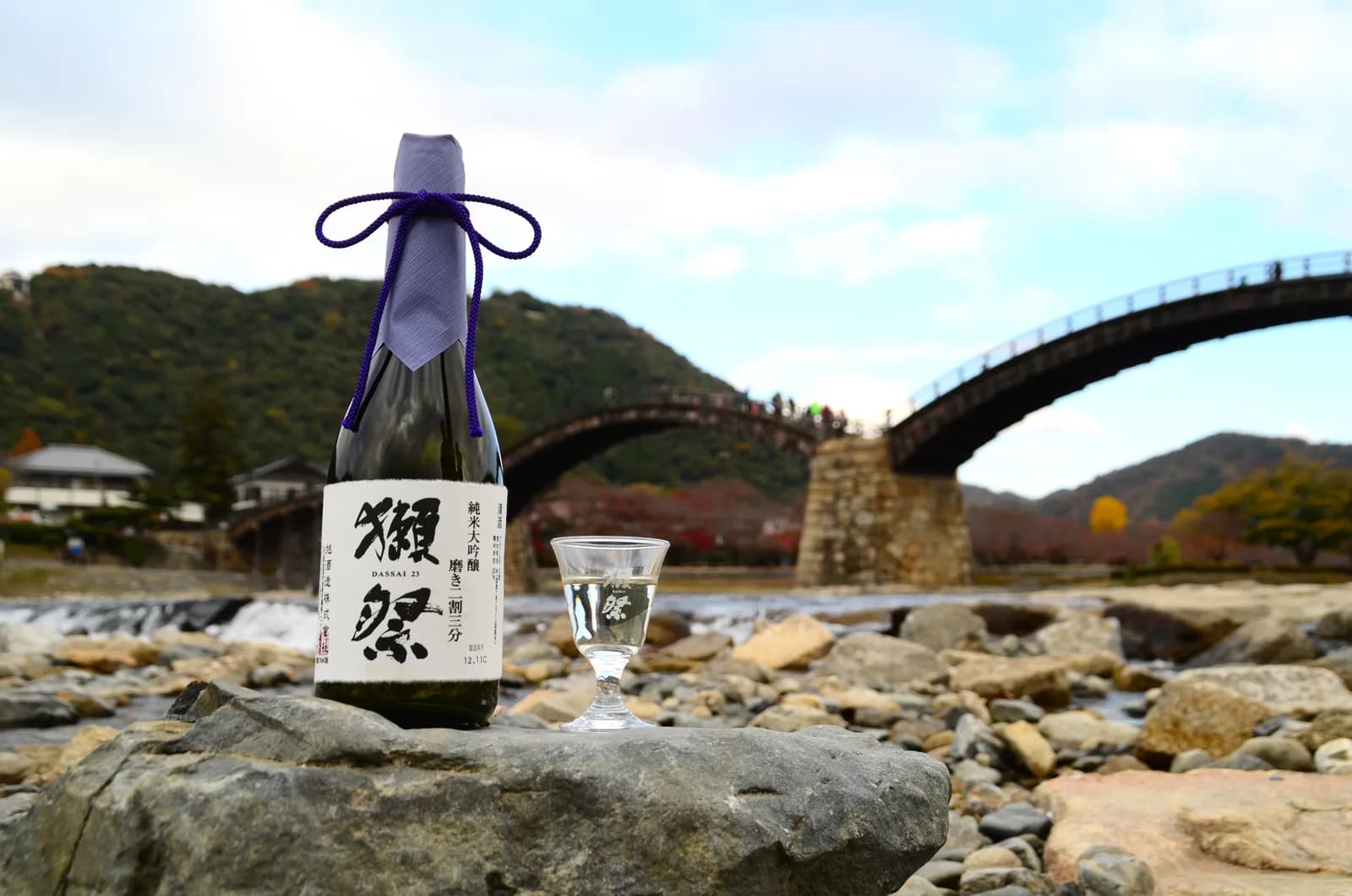 錦帯橋を背景に、白いラベルに「獺祭 磨き二割三分」と書かれた日本酒瓶が立っている