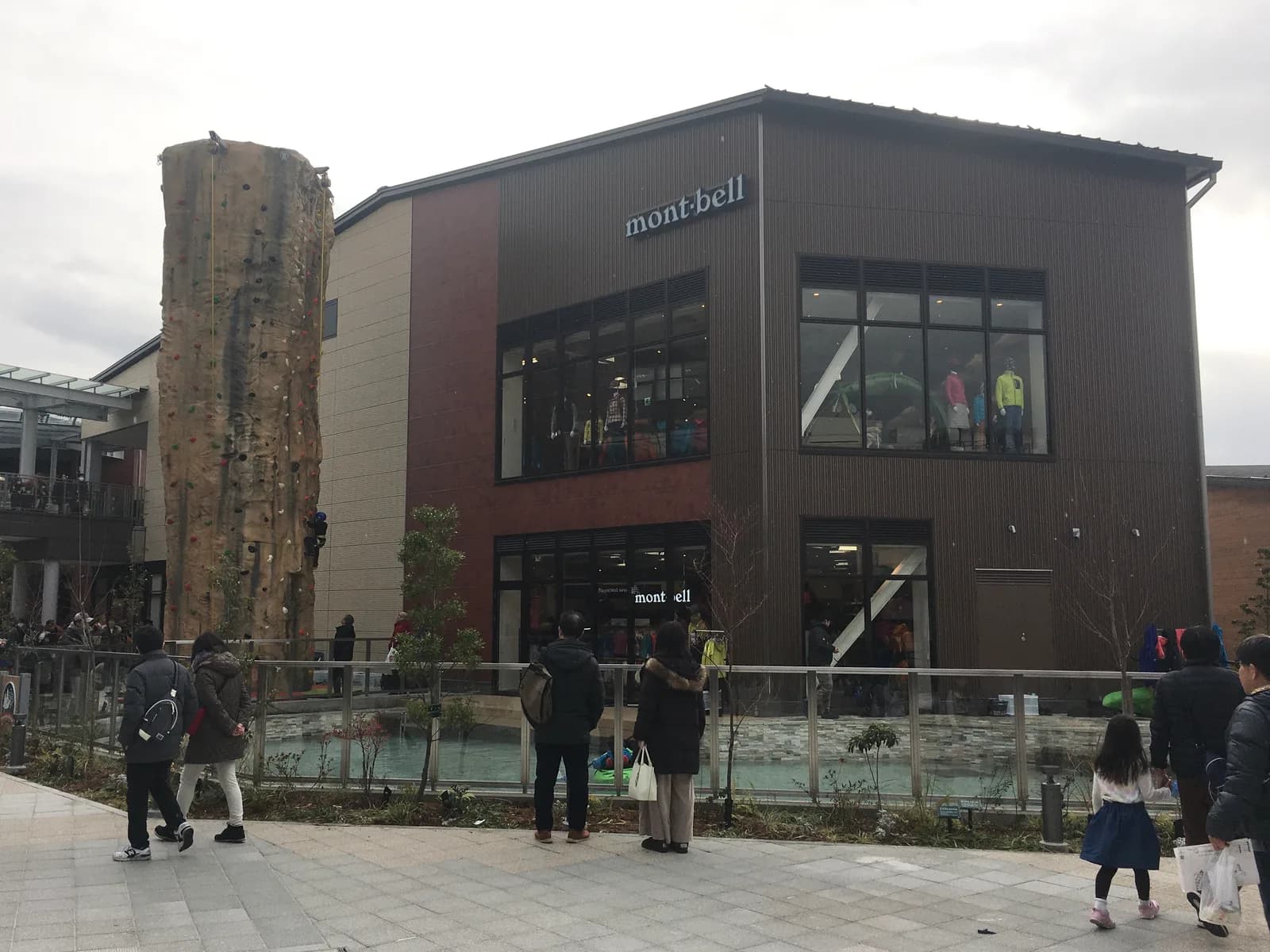 商業施設の一角に建つmont-bellの二階建て店舗。左手に人工の岩壁があり登っている人がいる。手前は人工池、家族連れが歩いている