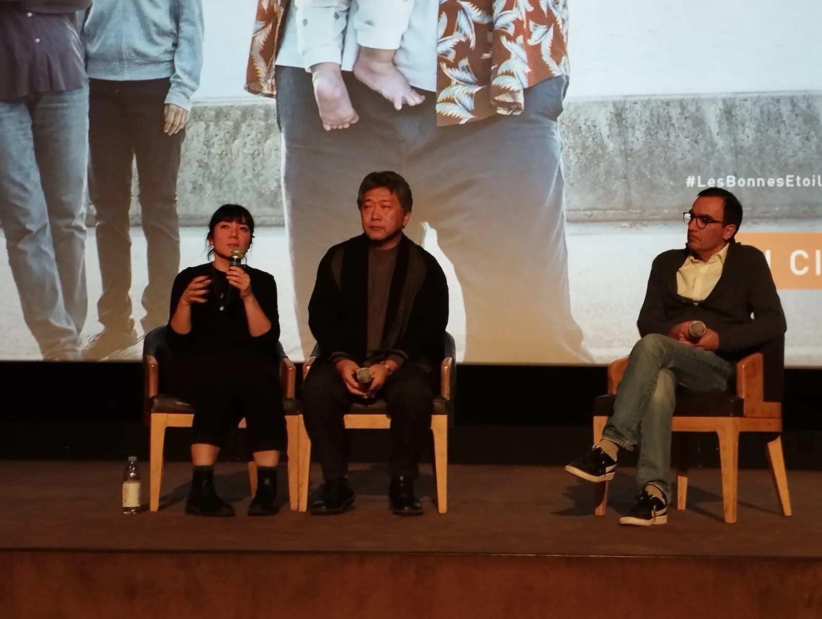 映画館の舞台上でマイクを持って話す男性。後ろにスクリーン