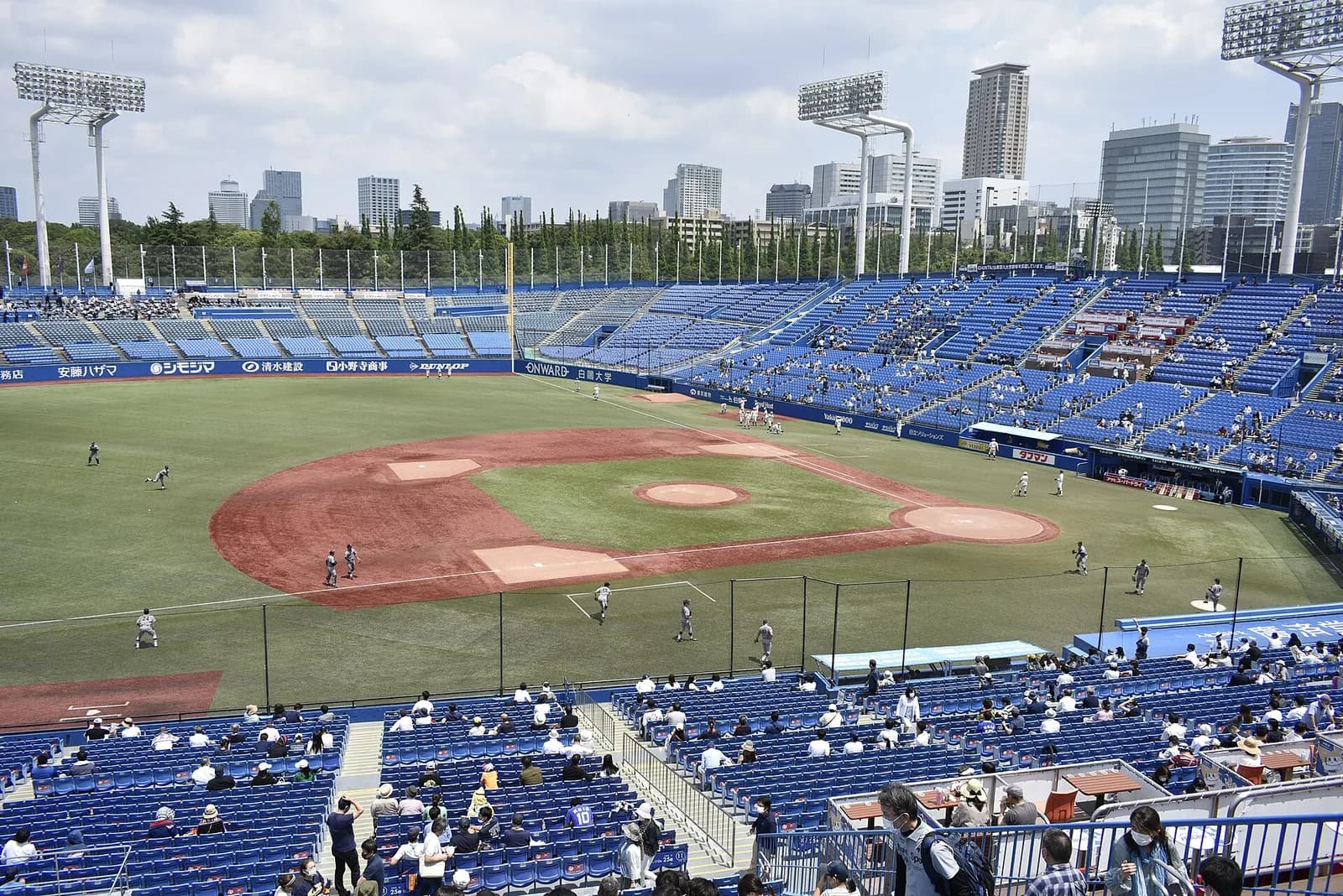 内野の上段から見下ろした球場。手前に青い座席と観客、奥にグラウンド全体と外野席、その向こうに都心のビル群が並ぶ