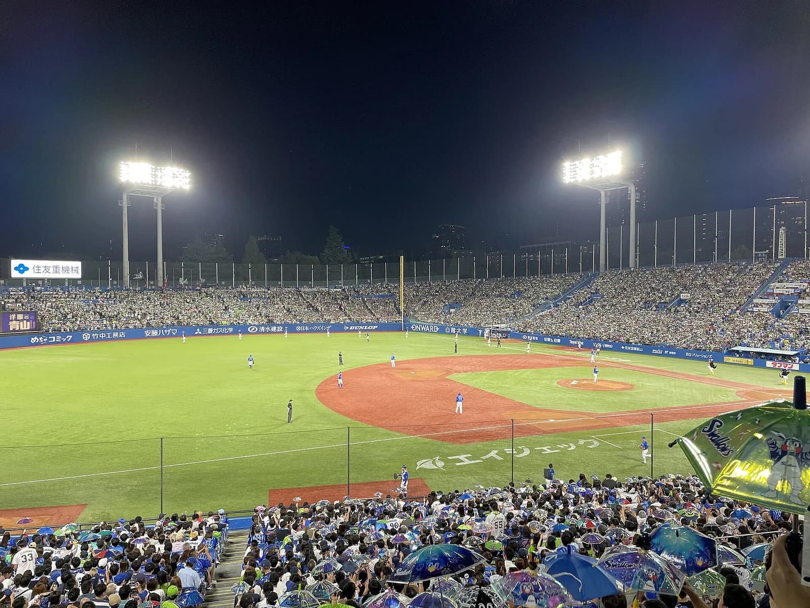 ナイターの神宮球場の全景。手前のスタンドで観客がチーム柄の小さな傘を掲げている