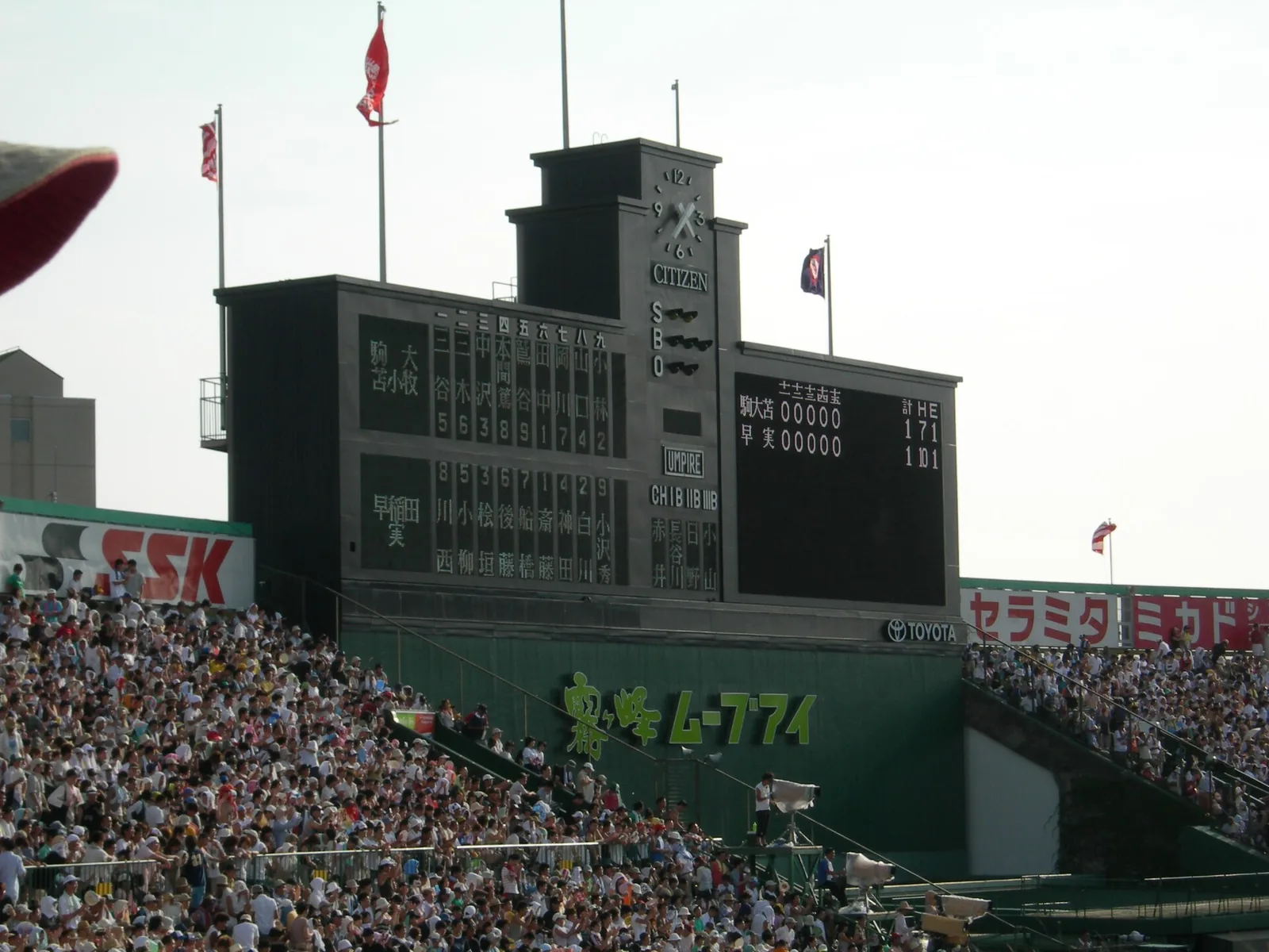 球場のスコアボード。両校の選手名が漢字で縦に並び、下のスタンドは観客で埋まっている