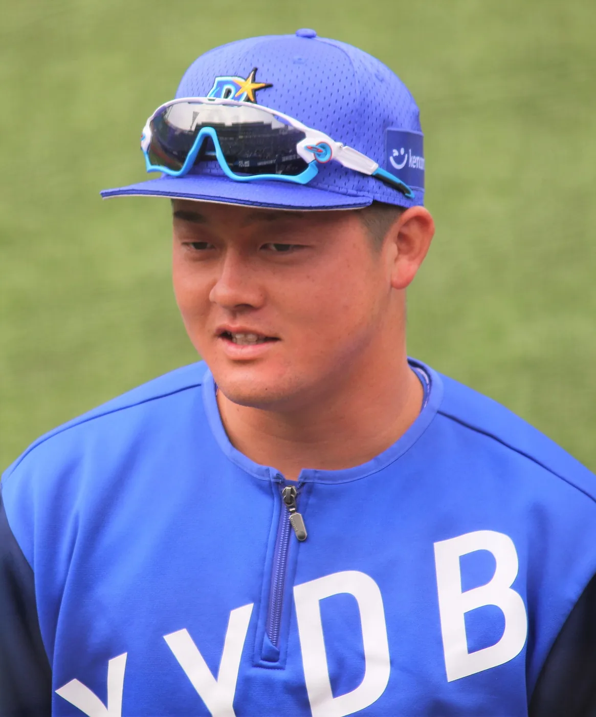 青い練習ウェアとキャップ姿の若い選手の上半身。帽子のつばにサングラスを載せている