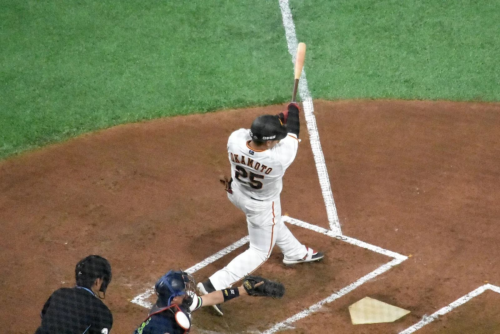 白いホームユニフォームの打者がスイングを振り抜いた直後を、スタンド上段から見下ろした構図。背中にOKAMOTOと背番号25