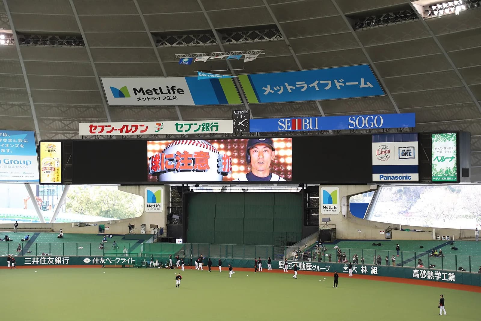 屋根の下のスコアボードと外野スタンド。左右の開口部から外の木々が見え、選手が試合前の練習をしている
