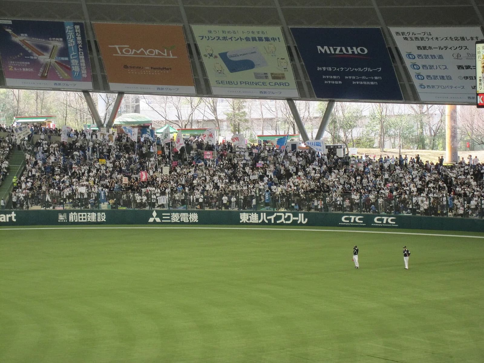 球場の外野席が観客で埋まり、旗や幟が何本も立っている。手前は外野の芝と守備位置につく外野手二人