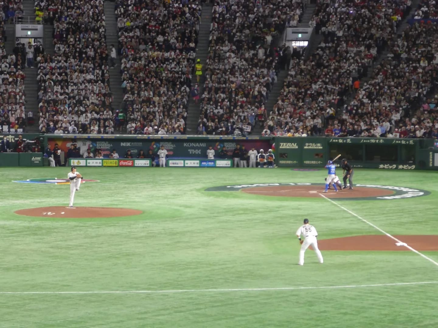 満員の屋根付き球場。マウンドで投球動作に入る投手と、打席の打者、内野の守備位置につく選手が見える