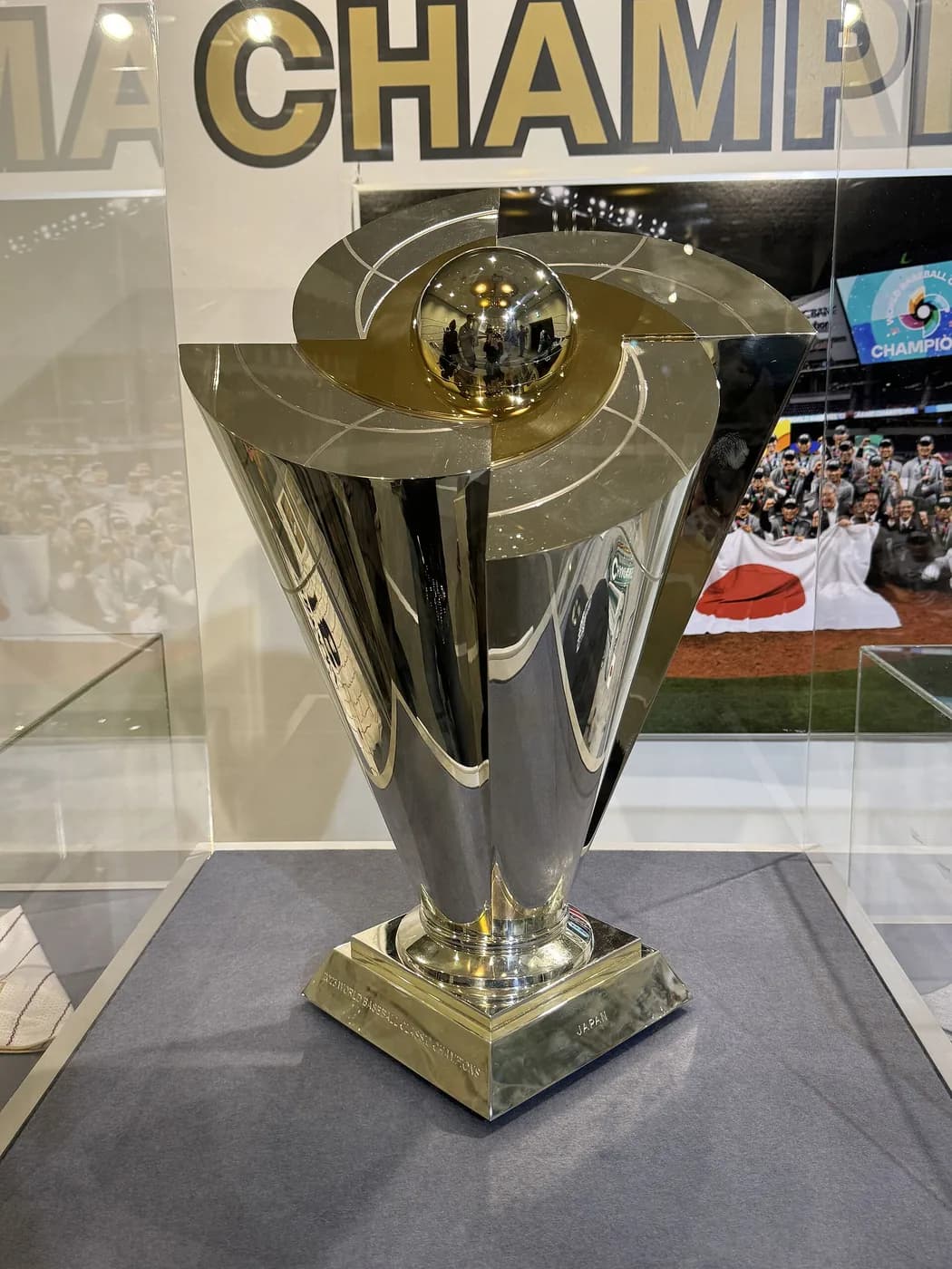 ガラスケースに展示された金属製のトロフィー。台座に文字が刻まれ、背景のパネルに優勝チームの集合写真がある