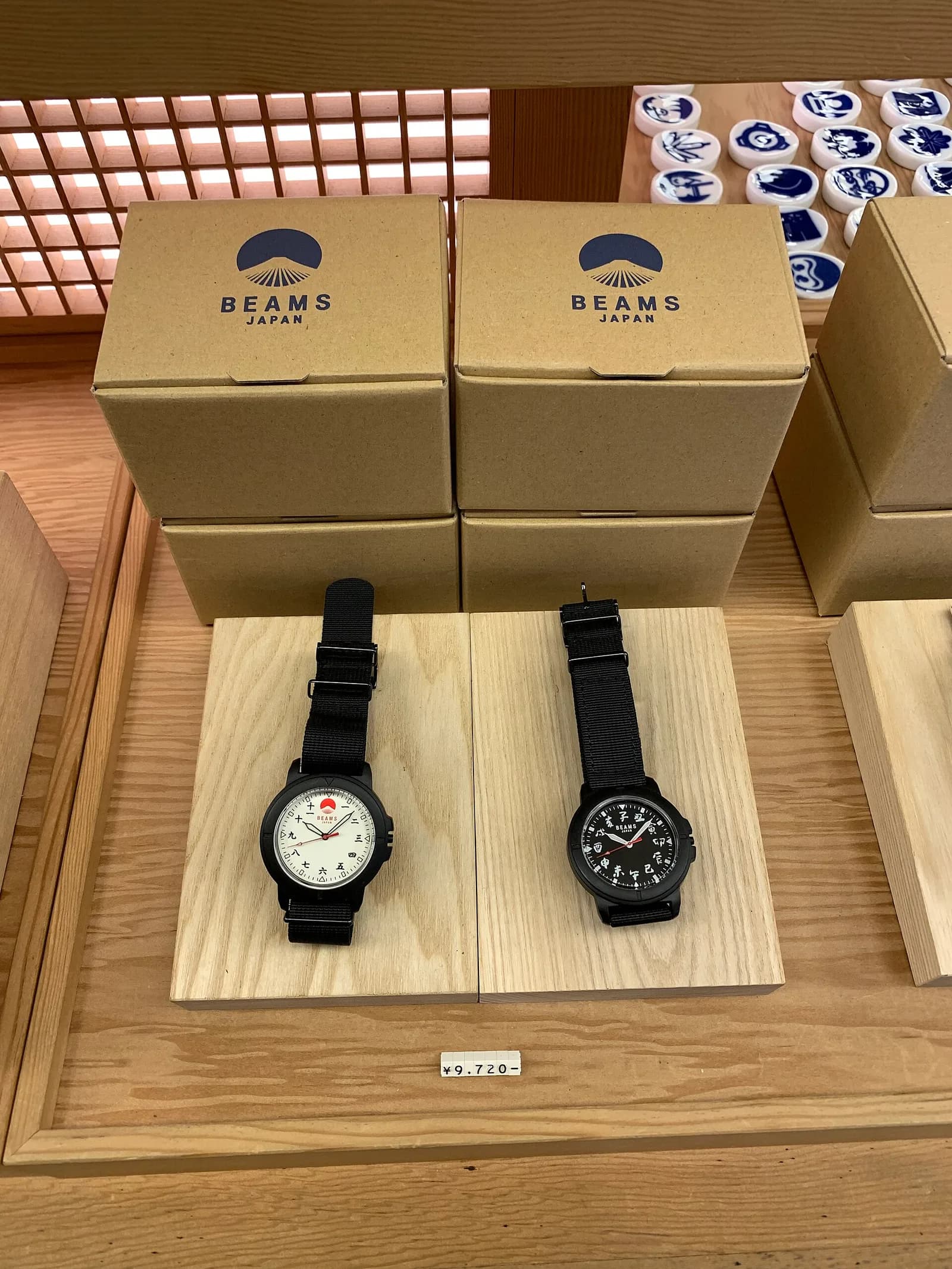 店の売り場で、木のブロックに載せられた二本の腕時計。後ろに商品の箱と値札が並ぶ