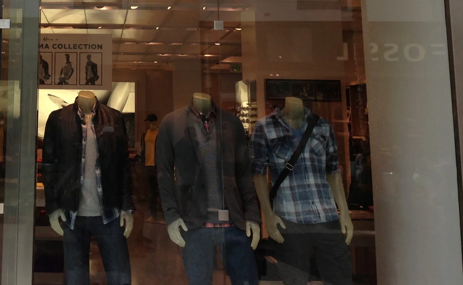 店頭のガラス越しに並ぶ三体のマネキン。焦茶の革調ブルゾンにグレーのTシャツとデニム、チャコールのニットジャケットに柄シャツ、青いチェックのシャツ。いずれも表にロゴは出ていない