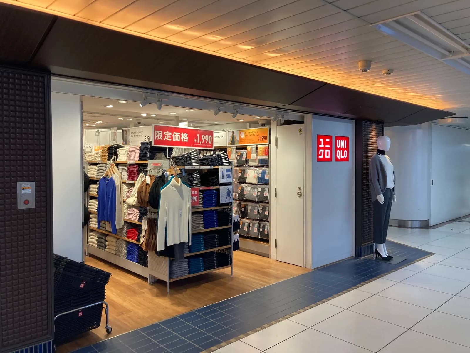 駅構内の小型店。畳んだニットが色ごとに積まれた棚と、下着のパックが並ぶ棚。手前に顔のないマネキンが一体立つ