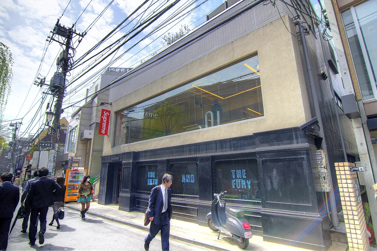 東京の通りに面したSupremeの店。赤い旗看板が出ていて、一階の窓にはTHE FILTH AND THE FURYという青いネオン文字。前の歩道を人が歩いている