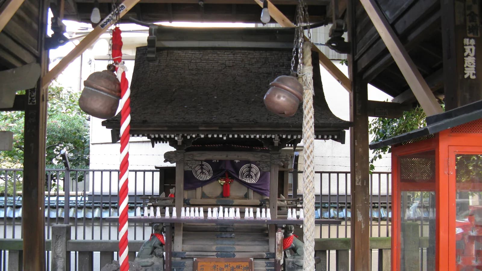 被官稲荷神社。木の覆屋の下に小さな社殿があり、左右に狐の像、正面に鈴と紅白の鈴緒が下がっている